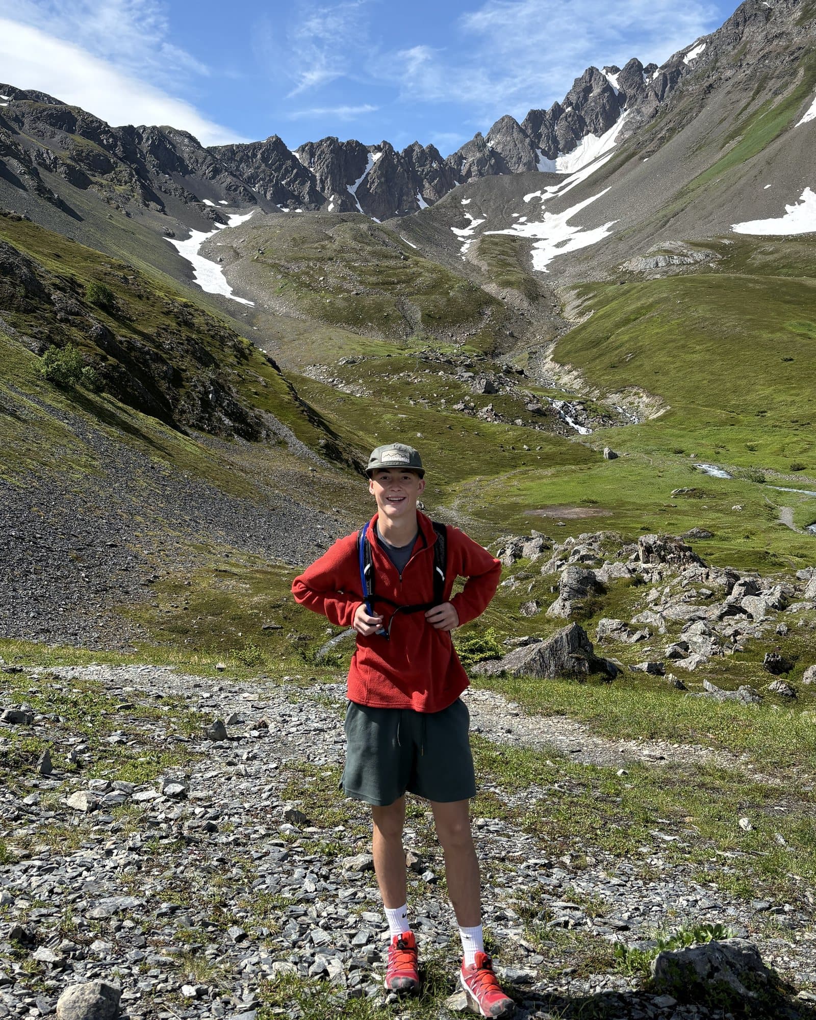Peter May standing on a gravel trail in a green mountain valley, snow still on the peaks behind him