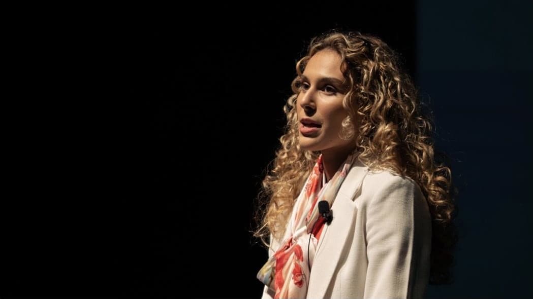 Speaker in a cream blazer lit by a single spotlight against a black stage backdrop