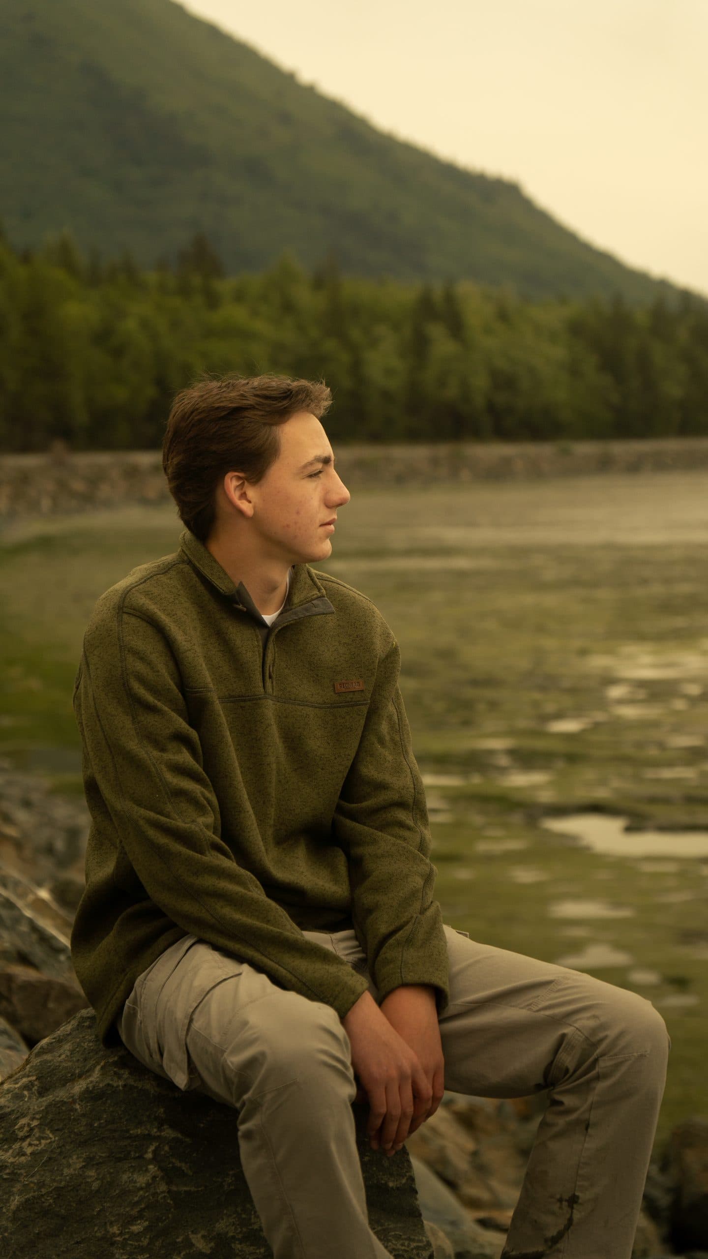 Senior in an olive fleece sitting on a boulder looking out across the tide flats, a forested mountain rising behind him