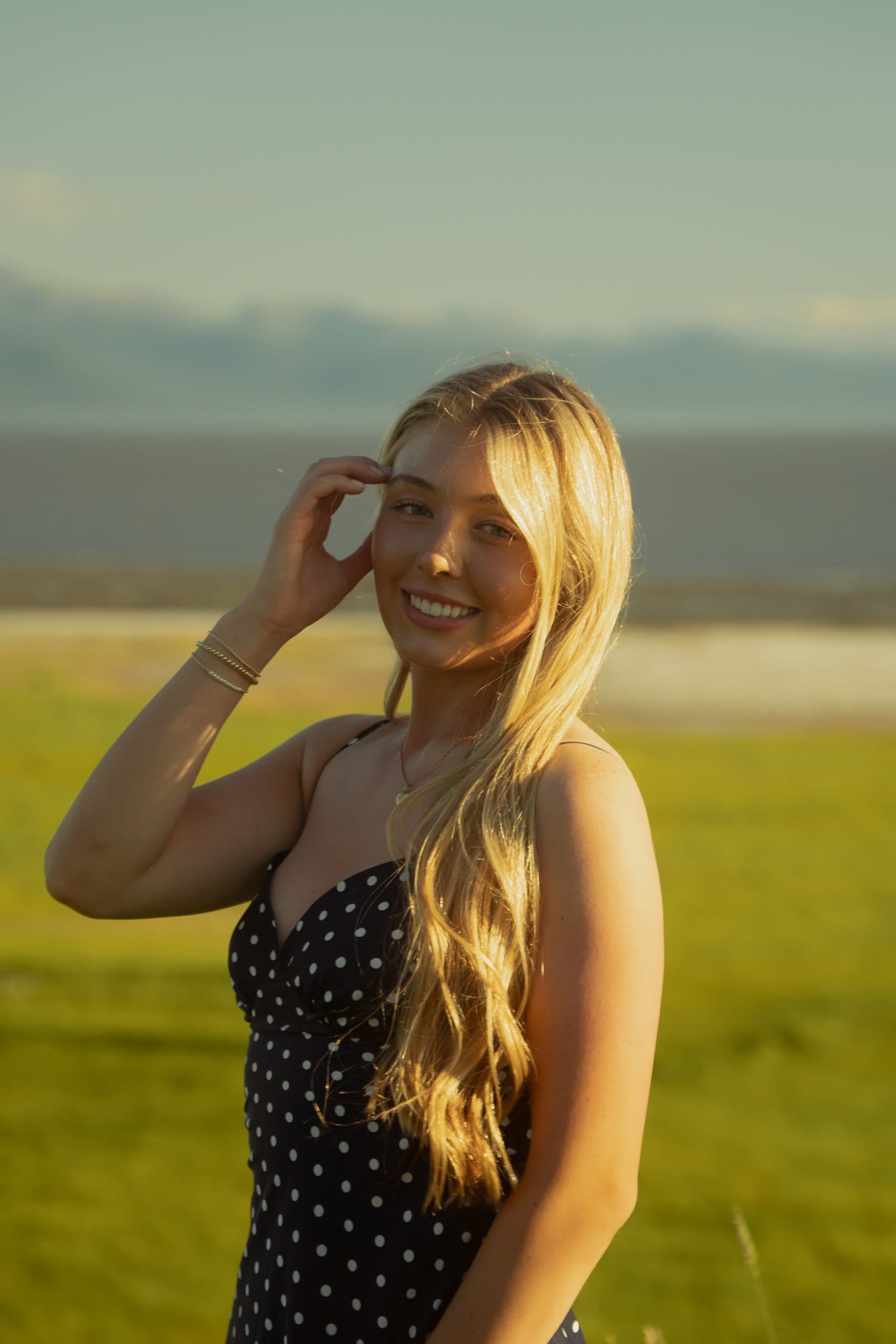 Senior in a polka-dot dress on a coastal bluff, brushing her hair back in low evening light