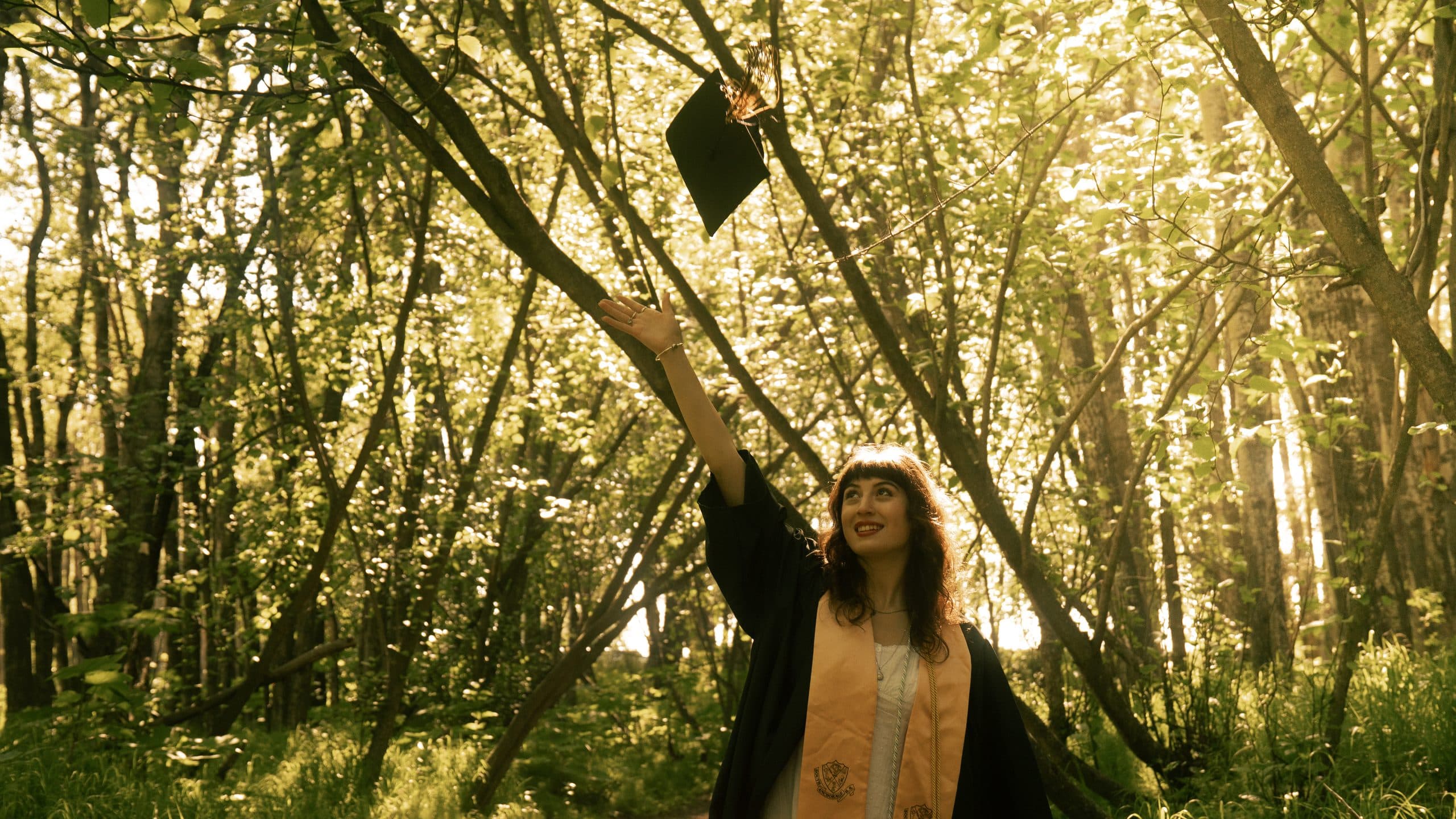 Graduate throwing her mortarboard into the air beneath tall sunlit summer trees