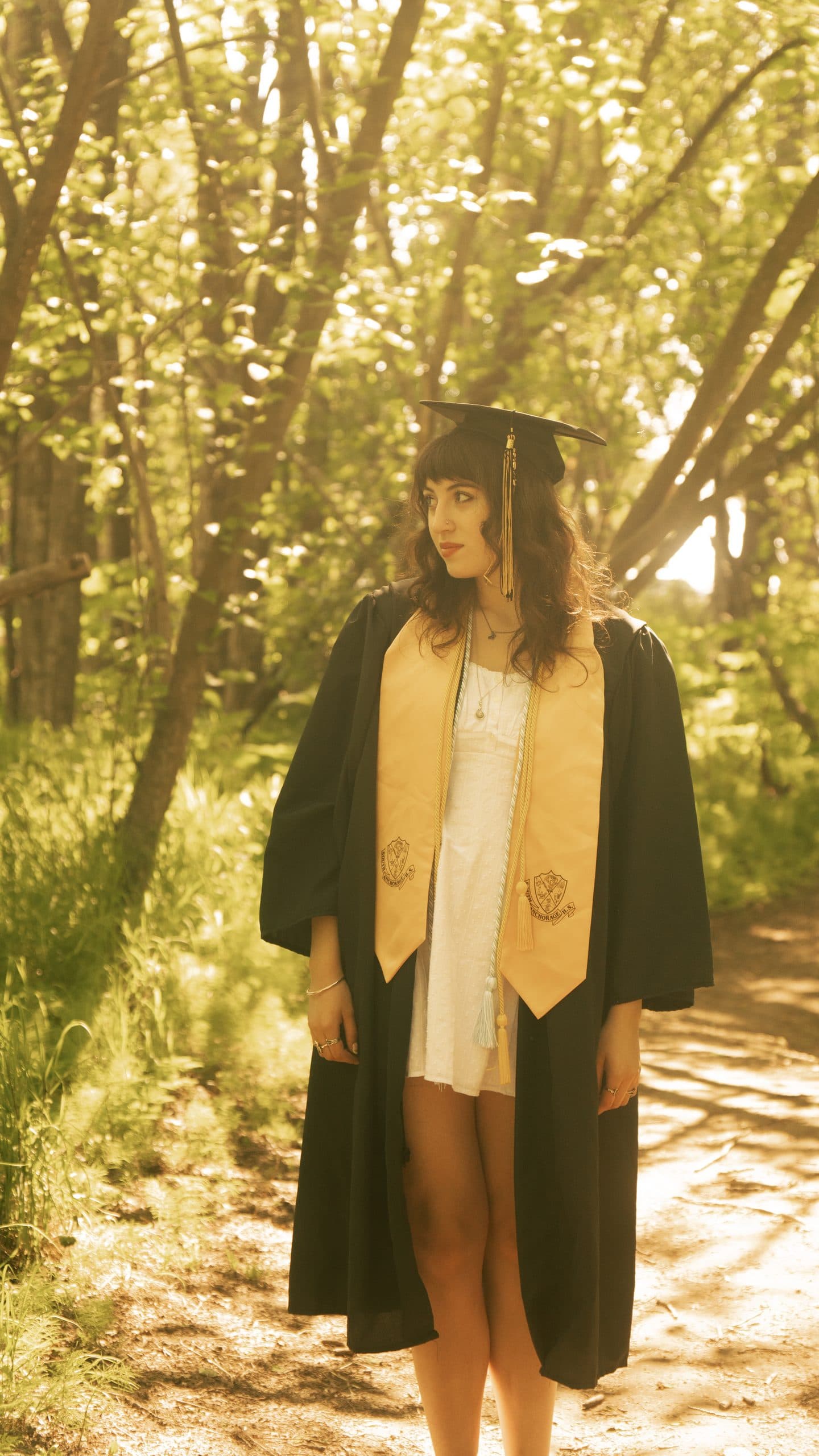 Graduate in cap, gown and gold honor stole standing on a sunlit forest path