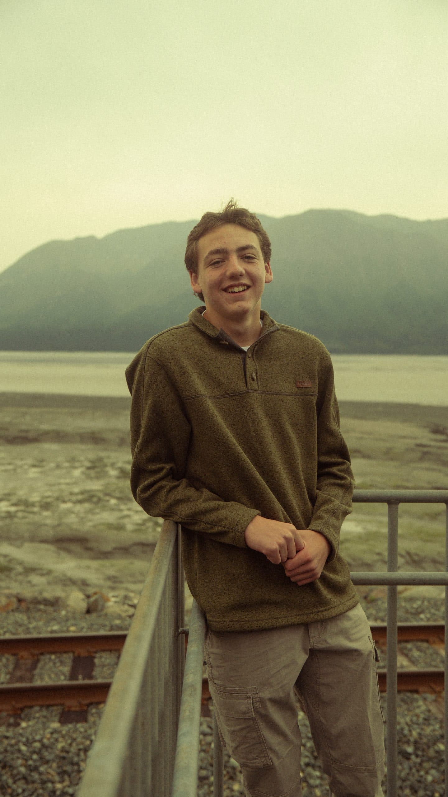 Senior in an olive fleece leaning on a metal railing above railroad tracks, the tide flats and a low mountain ridge behind him