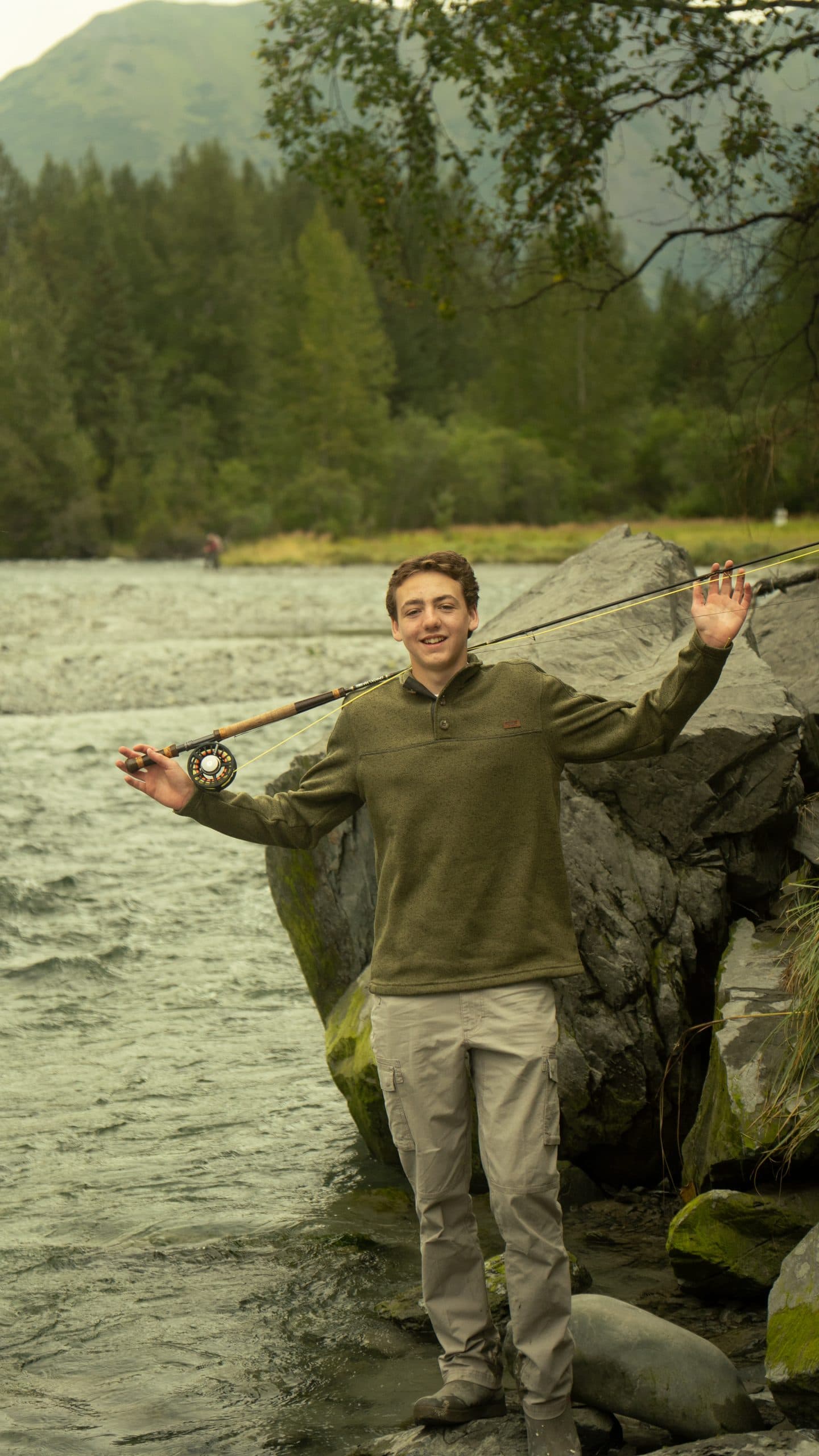 Senior in an olive fleece standing on a rocky riverbank with a fly rod across his shoulders, arms spread, spruce and a green ridge behind him