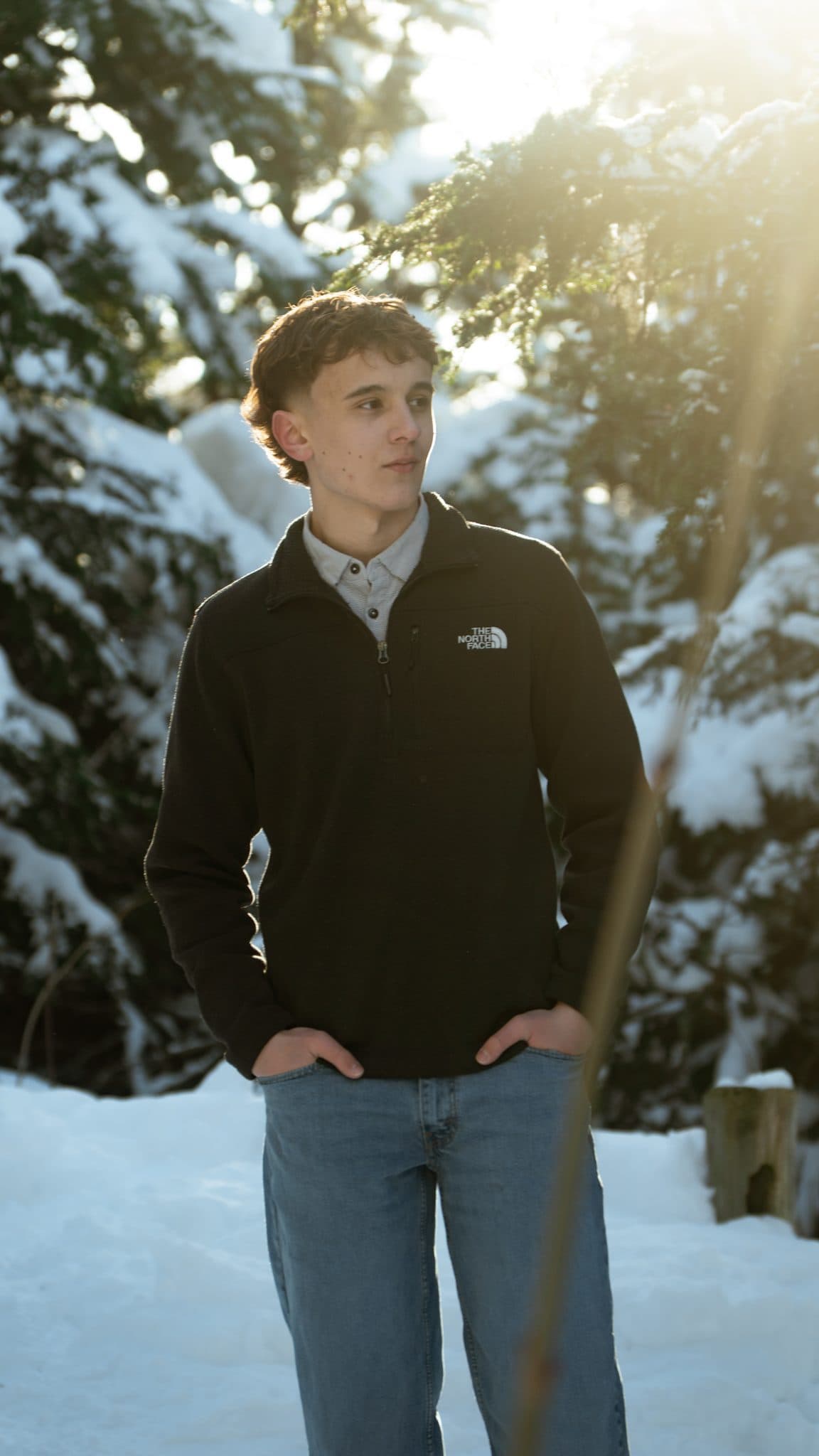 Senior in a dark fleece standing in deep snow, backlit by low winter sun through snow-covered spruce