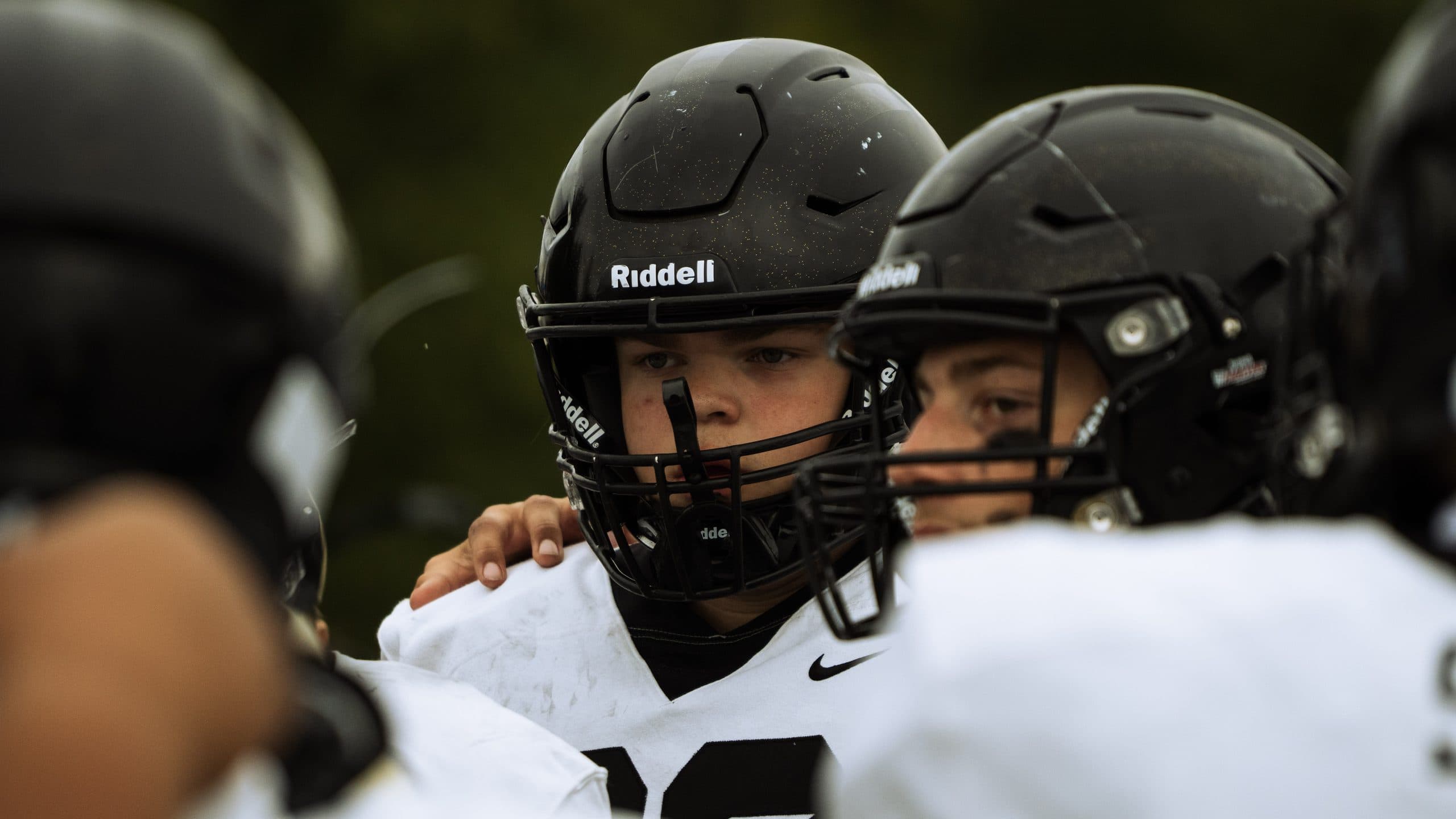Football player in a black helmet looking out from a tight cluster of teammates on the sideline, an arm across his shoulder
