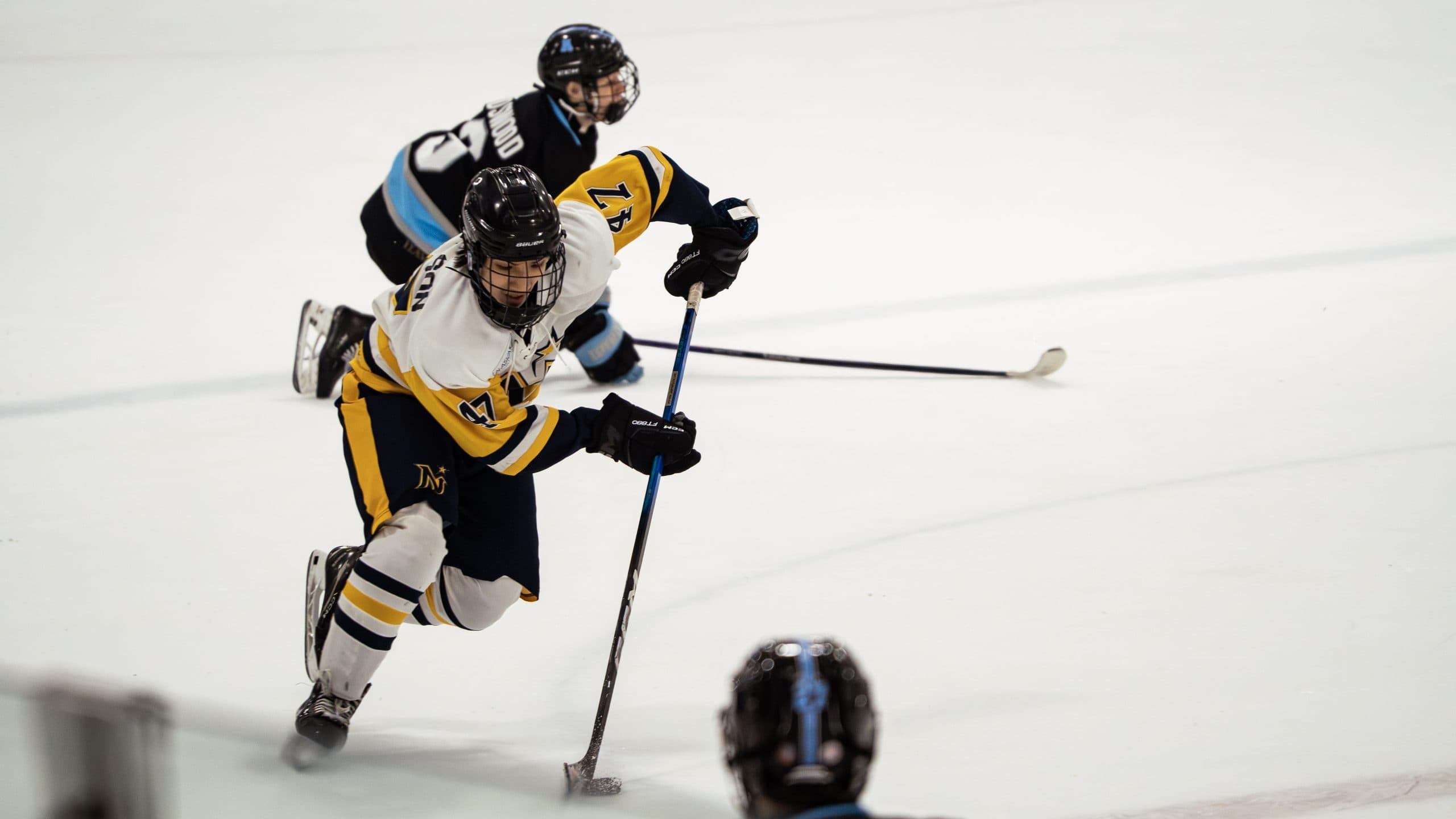 Hockey player in a white and gold jersey driving the puck up open ice, a defender in blue trailing behind him