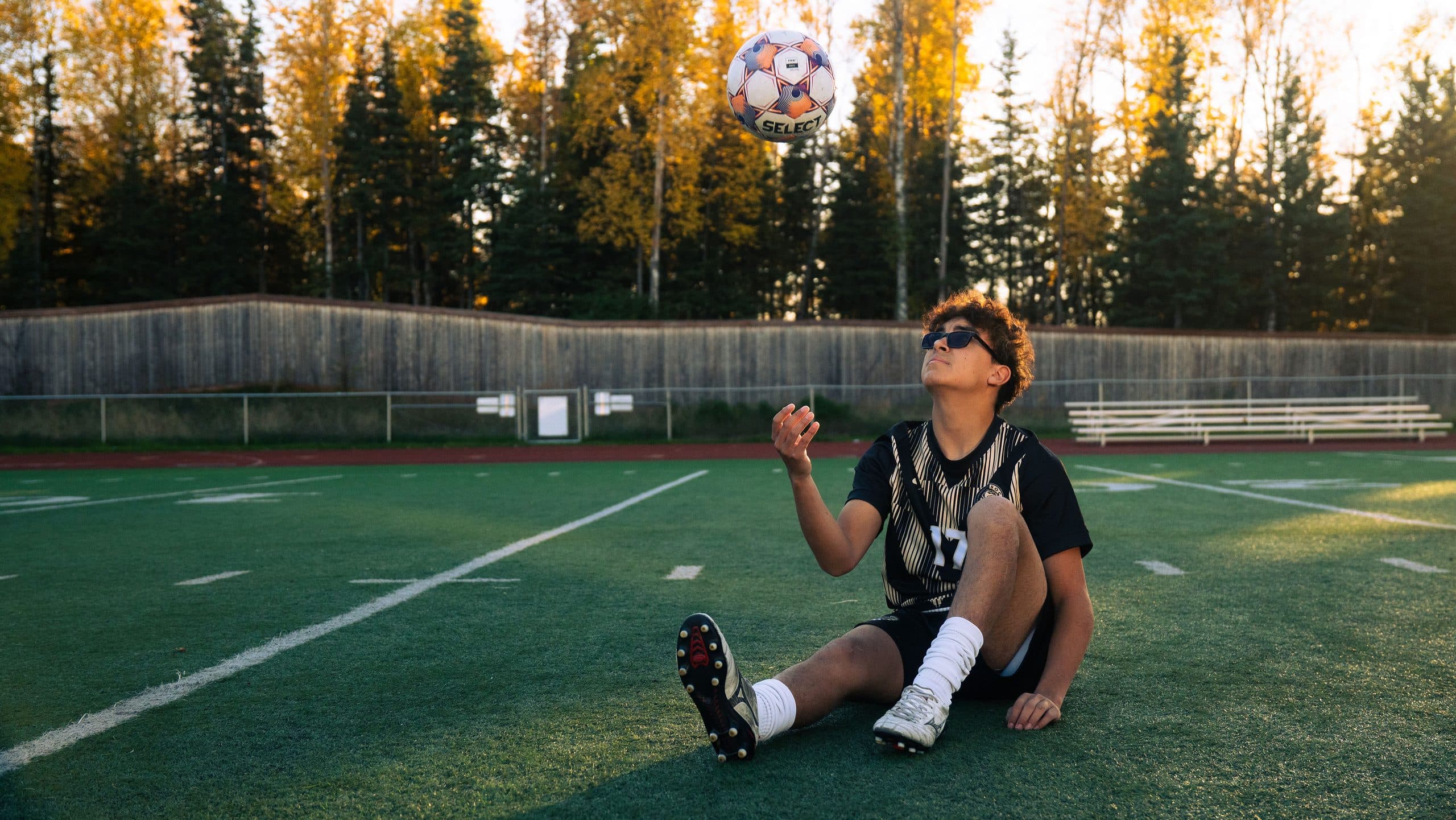 Soccer player sitting on the turf watching a ball spin in the air above him at golden hour