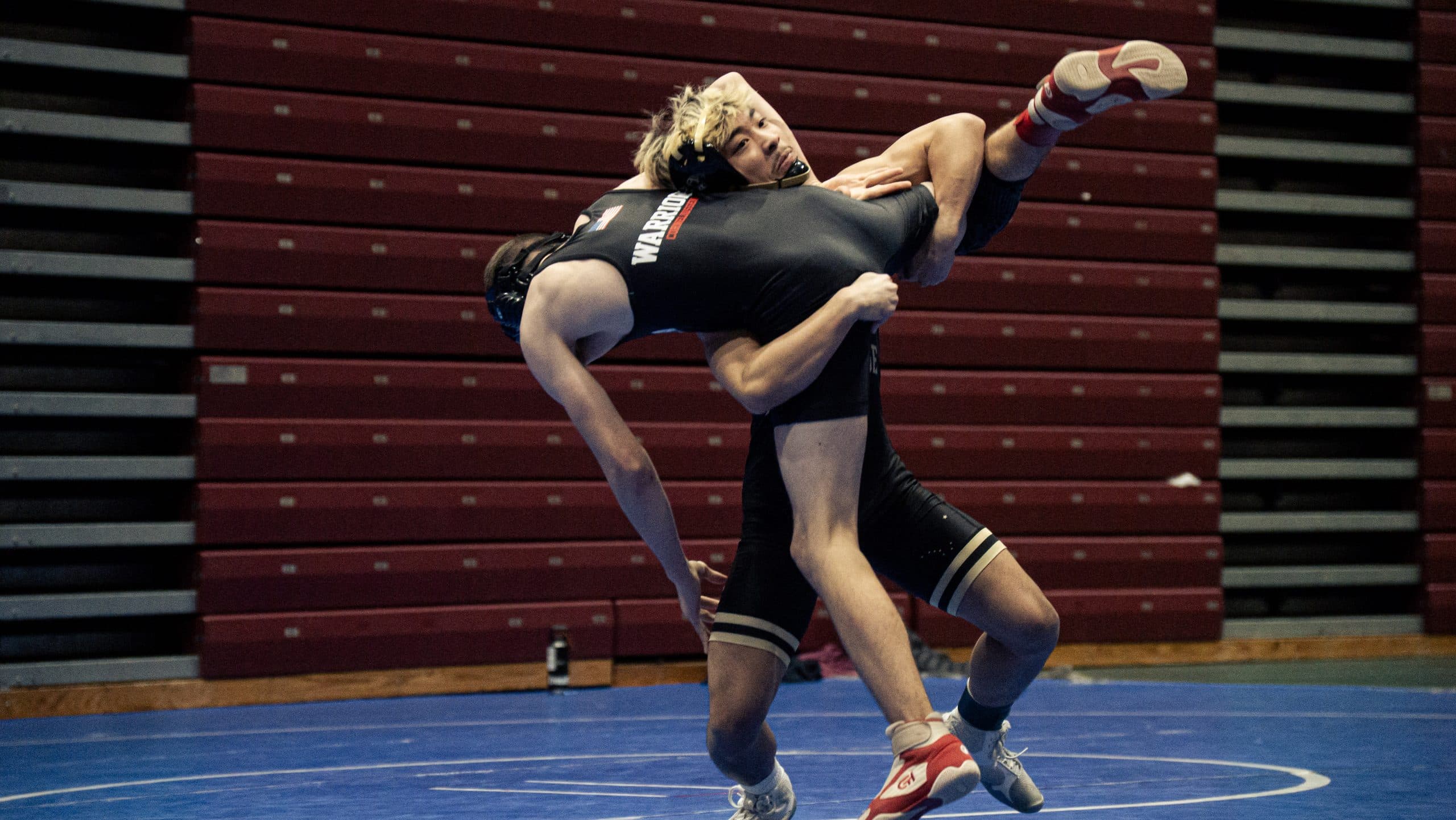 Wrestler lifting his opponent off the mat mid-throw in front of empty maroon bleachers